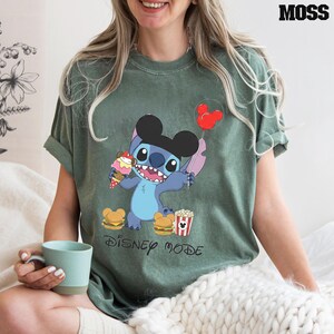 A woman wearing a green t-shirt with a cartoon character from Disney is sitting on a bed, holding a cup of coffee.