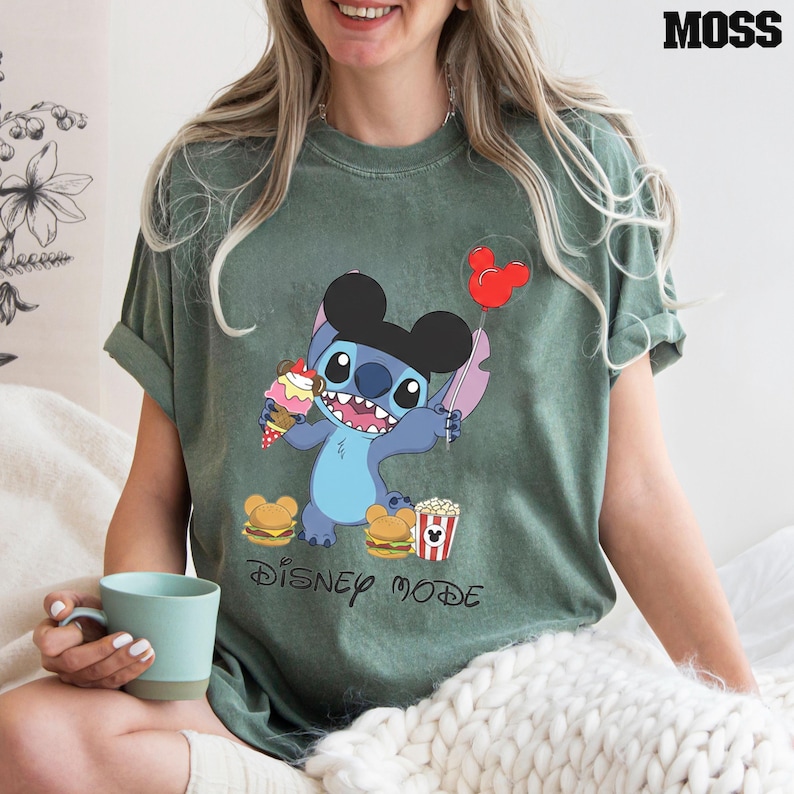 A woman wearing a green t-shirt with a cartoon character from Disney is sitting on a bed, holding a cup of coffee.