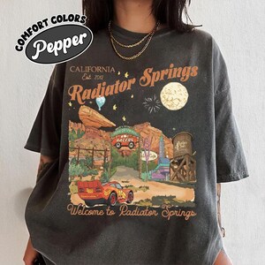 A person wearing a black t-shirt with a graphic design depicting a desert landscape with a car, a water tower, and a moon.