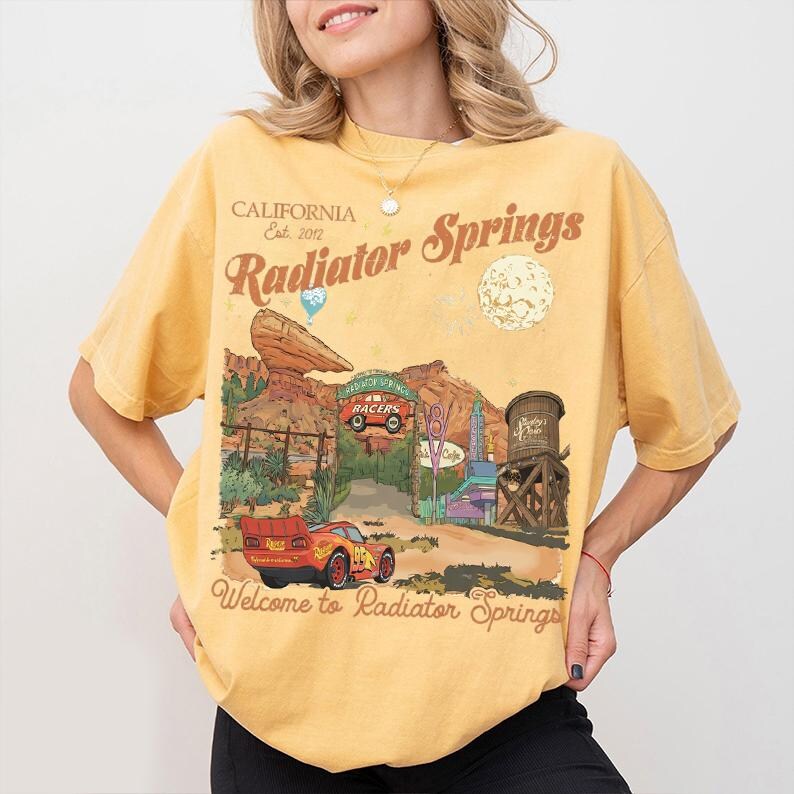 A woman wearing a yellow t-shirt with a graphic design depicting a scenic road leading to a car-themed location, along with the text "California Radiator Springs" and "Welcome to Radiator Springs".