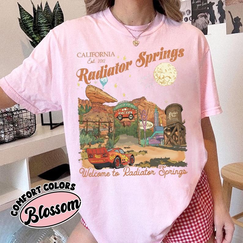 A person wearing a pink t-shirt with a graphic design depicting a desert landscape and various landmarks, including a car and a water tower, along with the text "California Radiator Springs Welcome to Radiator Springs".