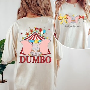 May include: Cream-colored t-shirt featuring a Dumbo graphic. The front of the shirt has a Dumbo design with the text "Don't just fly, soar." The back of the shirt has a circus tent design with Dumbo and the word "Dumbo" in red.