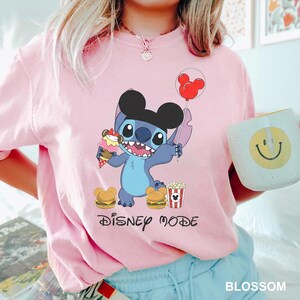A person wearing a pink t-shirt with a Disney character and the text "Disney mode" printed on it, holding a cup with a smiley face design.