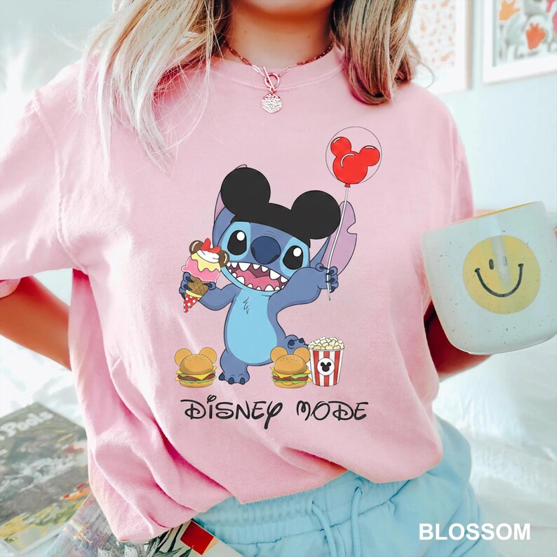 A person wearing a pink t-shirt with a Disney character and the text "Disney mode" printed on it, holding a cup with a smiley face design.