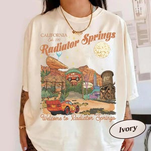 A person wearing a white t-shirt with a graphic design depicting a desert landscape with a car racing through it, along with a sign that says "Radiators Springs".