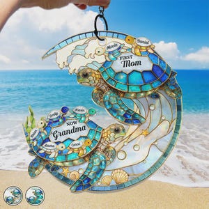 May include: A decorative, stained-glass style turtle family ornament. The ornament features two large turtles with smaller turtles labeled with names. The top turtle has the text "First Mom" and the bottom turtle has the text "Now Grandma". The ornament is in shades of blue and white.