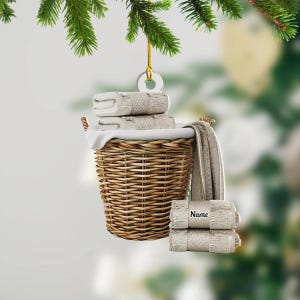 May include: A Christmas ornament featuring a woven wicker laundry basket filled with folded beige towels. A stack of towels hangs outside the basket, and another stack of towels with the word "Name" is below. The ornament hangs from a gold string.