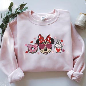 May include: A light pink sweatshirt featuring a Minnie Mouse graphic with heart-shaped sunglasses, a polka dot bow, a donut, and a cupcake. The design includes small red hearts. The sweatshirt has long sleeves and a crew neck.