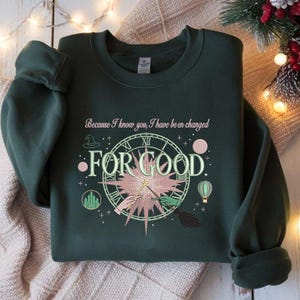 May include: Dark green sweatshirt featuring the phrase "Because I know you, I have been changed FOR GOOD". The design includes a clock, a witch's hat, a magic wand, and a broom, in a whimsical style.