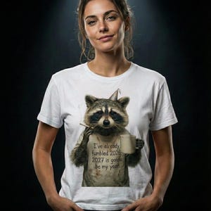 Può includere: T-shirt bianca con un procione con un cappello da festa, che tiene in mano una sigaretta e una tazza. Il testo sulla maglietta recita: "Ho già combinato un pasticcio nel 2026. Il 2027 sarà il mio anno."
