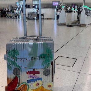 May include: A silver hard-shell rolling suitcase with an extendable handle, featuring a hand-painted design of tropical fruits, palm trees, disco balls, and signs reading "Just Married," "Bangkok," and "Malaysia" with the initials "SP" at the bottom.