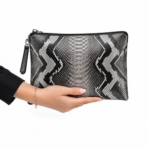 Pode incluir: Uma bolsa clutch retangular com um padrão de textura de pele de cobra em preto, prata e cinza com um design em ziguezague. A bolsa inclui um fecho de correr preto na borda superior com um puxador texturizado a condizer na lateral.