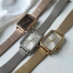 May include: Three rectangular wristwatches with mesh bands are displayed. The watches have different colored cases: rose gold with a black face, silver with a silver face, and gold with a gold face. The watch faces have the brand name "IEIE".