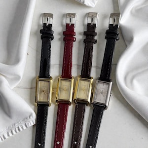 Pode incluir: Quatro relógios de pulso retangulares com pulseiras de couro nas cores preta, bordô e marrom. Os mostradores dos relógios são dourados e prateados, com a palavra "So Cozy" impressa no mostrador. Os relógios são exibidos em uma superfície branca.