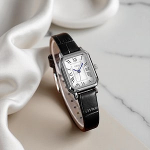 Puede incluir: Un reloj rectangular plateado con esfera blanca y correa de cuero negro. La esfera del reloj presenta números romanos y la marca "GEDI". El reloj tiene un diseño clásico.