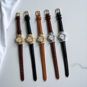 Könnte beinhalten: Fünf Armbanduhren mit Lederarmbändern in verschiedenen Farben: Dunkelbraun, Schwarz, Beige und zwei Brauntönen. Die Zifferblätter sind rund mit römischen Ziffern. Die Gehäuse sind gold- und silberfarben.