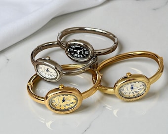 May include: Four oval-faced bangle watches in gold and silver tones. The watch faces display Roman numerals and hands. One watch has a black face, while the others have white or gold faces. The watches are displayed on a white surface.