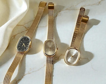 May include: Three gold-toned wristwatches with oval faces and mesh bands are displayed. The watch faces are in black, gold, and silver. The watches are arranged on a marble surface with a white fabric in the background.