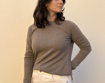 May include: A long-sleeved, crew-neck sweater in a solid taupe-brown color, paired with high-waisted, off-white denim jeans featuring a visible metal button closure at the waist and classic five-pocket styling.