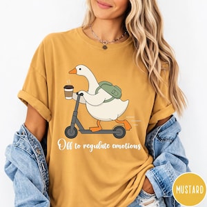 Puede incluir: Camiseta de color mostaza con un dibujo de un ganso de dibujos animados montado en un patinete, sosteniendo una taza de café. El ganso lleva una mochila y el texto "Off to regulate emotions" está impreso debajo. La camiseta tiene las mangas dobladas.