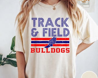 May include: Ivory-colored t-shirt with the words "TRACK & FIELD BULLDOGS" in blue and red lettering. The design includes a winged foot graphic, a symbol of track and field.