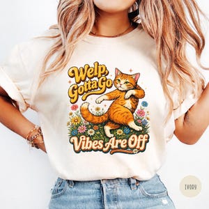 Puede incluir: Camiseta color marfil con un gato atigrado naranja de dibujos animados caminando por un campo de flores. El texto "Welp Gotta Go Vibes Are Off" está impreso encima y debajo del gato en una fuente retro.
