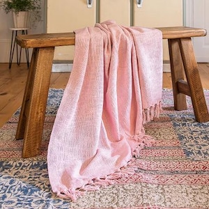 May include: A pink woven throw with a fringed edge is draped over a wooden bench. The bench rests on a patterned rug with blue, red, and beige sections. A vintage kitchen cabinet is in the background.