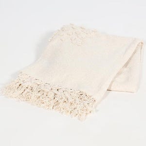 May include: Cream-coloured throw blanket with a textured weave and decorative fringe along one edge. The blanket features a subtle floral pattern near the top. The soft, neutral tones make it a versatile home decor item.