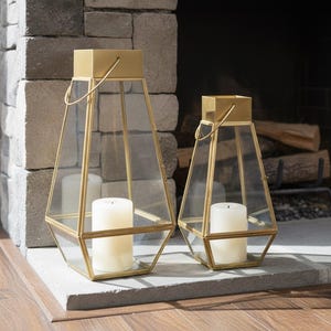 May include: Two geometric lanterns with gold-coloured metal frames and clear glass panels. Each lantern holds a white pillar candle. The lanterns are set on a stone surface in front of a stone fireplace. The frames have a handle.