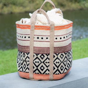 May include: A cylindrical woven basket with two tan fabric handles, featuring horizontal bands of orange, cream, brown, and a black diamond pattern. The basket is filled with a cream-coloured fabric, showcasing a textured weave throughout the exterior.