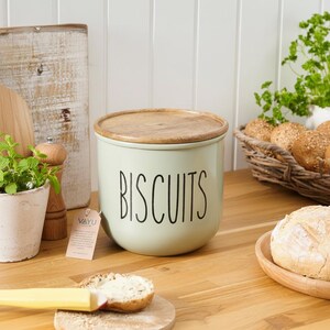 May include: A pale green biscuit jar with a wooden lid, labelled "BISCUITS" in black. The jar is on a wooden surface. A small plant in a pot and a basket of bread rolls are in the background.