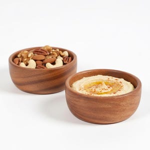 May include: Two wooden bowls on a white background. One bowl contains a mix of nuts, including walnuts, pecans, almonds, and cashews. The other bowl holds hummus, drizzled with oil and sprinkled with paprika.