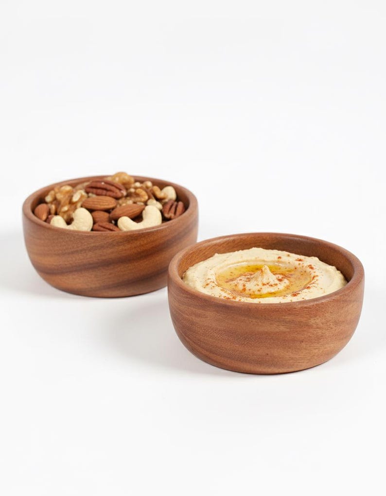 May include: Two wooden bowls on a white background. One bowl contains a mix of nuts, including walnuts, pecans, almonds, and cashews. The other bowl holds hummus, drizzled with oil and sprinkled with paprika.