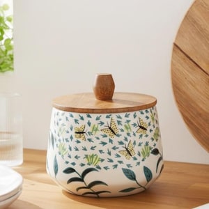 May include: A white ceramic canister with a tapered shape, featuring a pattern of yellow butterflies, green leaves, and small teal arrows. The container has a round, flat wooden lid with a small, central, faceted wooden knob handle on top.