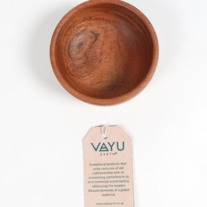 May include: A small, round wooden bowl with a rich brown color and visible wood grain. The bowl is empty and sits on a white surface. A tag with the text "VAYU EARTH" is below the bowl.