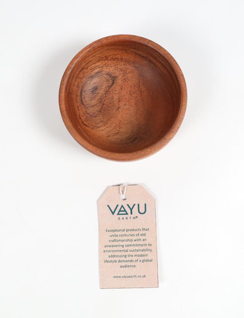 May include: A small, round wooden bowl with a rich brown color and visible wood grain. The bowl is empty and sits on a white surface. A tag with the text "VAYU EARTH" is below the bowl.