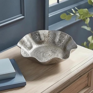May include: A silver-toned metal bowl with a scalloped, wavy rim and a textured surface featuring a repeating chevron or herringbone pattern. The bowl has a shallow, wide basin and a metallic finish that reflects light across its intricate design.