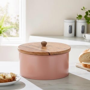 May include: A cylindrical storage container with a matte dusty pink metal body and a circular wooden lid featuring a small, centred wooden knob handle. The container has a smooth, solid-coloured finish and a natural wood grain pattern on the lid.