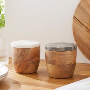 May include: Two wooden containers with marble lids. One has a white lid, the other a grey lid. The containers are cylindrical with a natural wood grain finish. The items are on a wooden surface, suggesting kitchen or home decor.