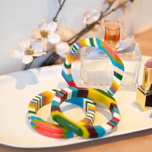 May include: Three colorful, geometric bangles with a striped design in shades of red, yellow, green, blue, and white. The bangles are arranged on a white tray with a perfume bottle, lipstick, and floral accents.