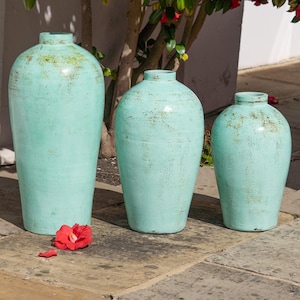 May include: Three turquoise ceramic vases of varying sizes. The vases have a distressed finish with hints of green and brown. The tallest vase is approximately 76 cm tall. A red flower lies on the stone surface in front of the vases.