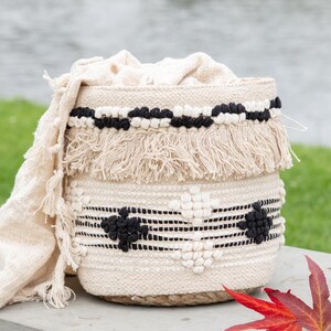 May include: A cylindrical woven basket featuring a cream-coloured base with black and white tufted geometric patterns and fringe detailing. A matching cream-coloured fabric cloth is draped over the side of the basket, which has a natural jute base.