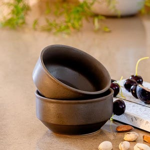 May include: Two stacked dark brown ceramic bowls with a matte finish and a wide, circular rim. The bowls feature a simple, functional design with a slightly tapered base. The dark, earthy tone of the ceramic contrasts with the smooth, rounded edges.