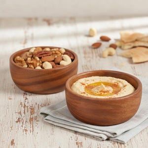 May include: Two wooden bowls on a white, distressed surface. One bowl contains a mix of nuts, including walnuts, pecans, and cashews. The other bowl holds hummus, garnished with oil and paprika. A gray napkin is under the hummus bowl.