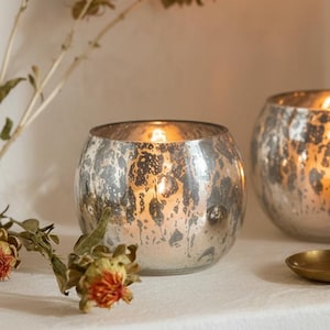 May include: Two silver mercury glass candle holders with lit candles. The round votives have a distressed finish and sit on a white surface. Dried flowers and a small gold dish are also present.