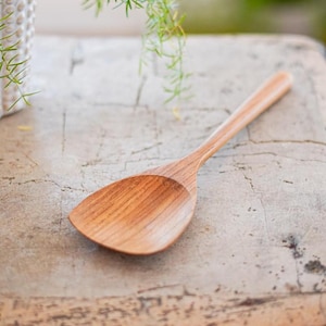 May include: A light brown wooden spoon with a wide, flat-edged bowl and a long, slender handle. The spoon features a visible natural wood grain pattern throughout its surface, showcasing a smooth, polished finish and a curved, functional design.