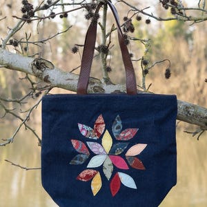 May include: A navy blue tote bag with brown leather handles and a colourful floral appliqué. The flower design features various patterned fabric petals in shades of red, pink, yellow, and blue. The bag is hanging from a tree branch.