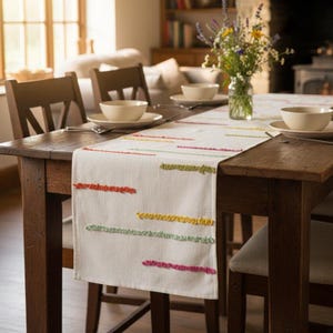 May include: A cream-coloured table runner featuring horizontal tufted lines in vibrant shades of orange, yellow, green, and pink. The runner is laid across a dark wooden dining table set with white bowls, silverware, and a glass vase of flowers.