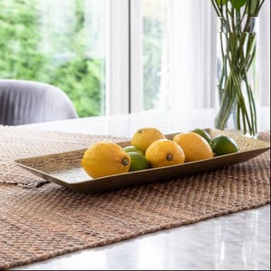 May include: A gold-coloured rectangular decorative tray holding lemons and limes. The tray is on a woven runner on a table. A clear glass vase with green stems is in the background, near a window.