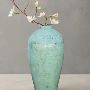 May include: A turquoise ceramic vase with a distressed finish, holding a branch of white flowers. The vase has a rounded body and a narrow neck. The flowers have delicate white petals. A decorative home accent, the vase is a stylish addition to any room.